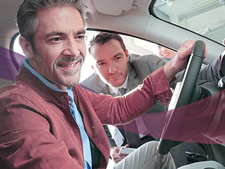 A customer checks out a car from inside while the salesman peeps inside through the window