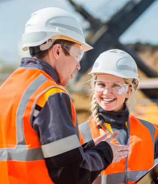Two workers wearing safety gear, in a friendly discussion, at an outdoor location, with heavy machinery in the background