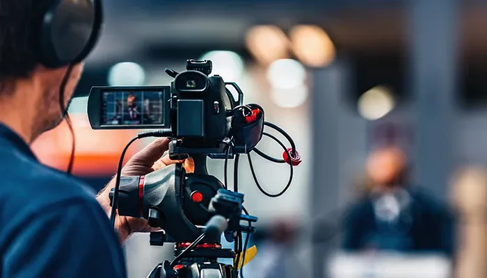 A man holds a video camera, focused on recording a video in an outdoor setting.