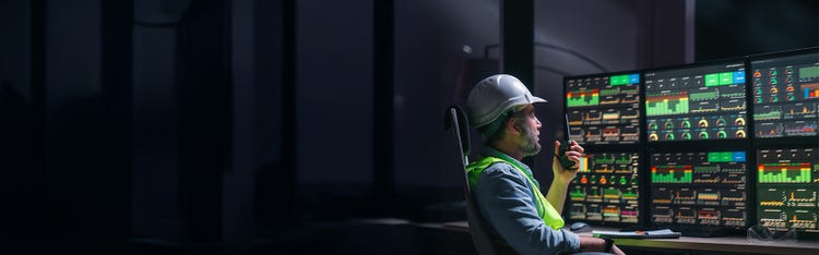 A male worker in hard hat and safety vest sitting in front of multiple computer monitors.