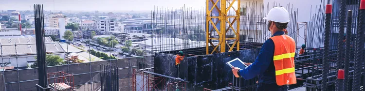 A construction worker wearing a hard hat and safety vest, actively working on a construction site with machinery in the background.