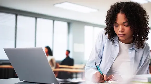 A woman writing down notes with her laptop open.