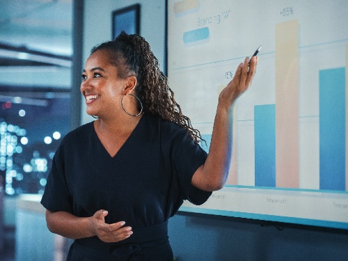 A woman pointing to a monitor screen while presenting at workplace