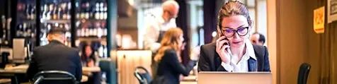 A woman talking on her cell phone while working on a laptop, against a blurred out background of a cafeteria