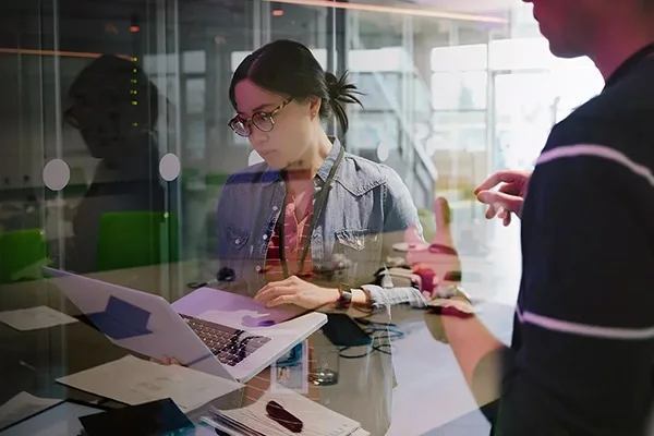 Two individuals collaborating at a laptop in a modern office setting, focused on their work.