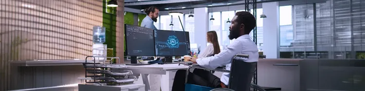 Man in a wheelchair works at a desk with dual monitors in a modern office; two colleagues are talking in the background.