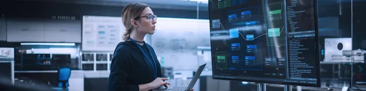 A female engineer holding a laptop is looking at a digital screen showing process data