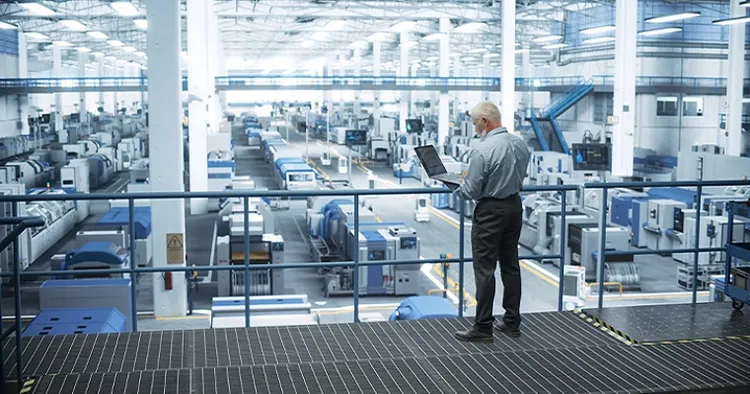 Person standing on a factory mezzanine using a laptop above an automated production floor.