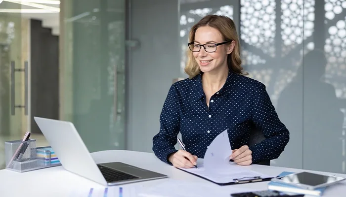 Individual with glasses working on laptop reviewing documents in office