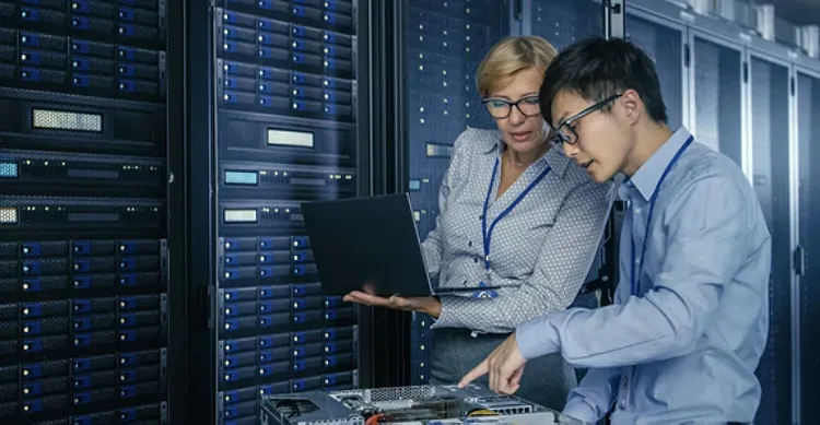 Two IT professionals in a server room, examining equipment. One holds a laptop, discussing data. The scene conveys focus and teamwork.