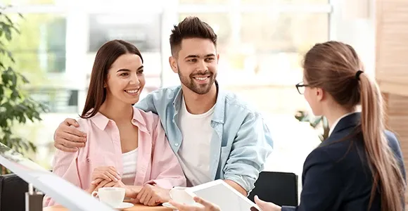 A smiling couple sits across a desk from a businesswoman in an office setting. The couple appears engaged and happy, suggesting a positive interaction.