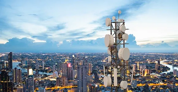 A tall telecommunications tower with numerous antennae overlooks a sprawling cityscape at dusk. The sky is clear, with a soft blue hue and scattered clouds.