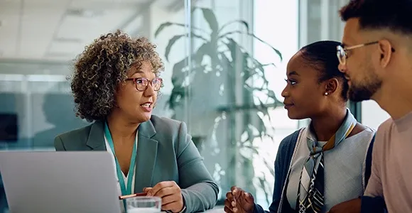 A professional woman with curly hair speaks to two colleagues at a table. They appear engaged and attentive, sitting in an office setting with natural light.