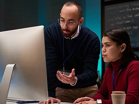 A woman sitting in front of a computer, while a man standing next to her is looking at the screen and talking